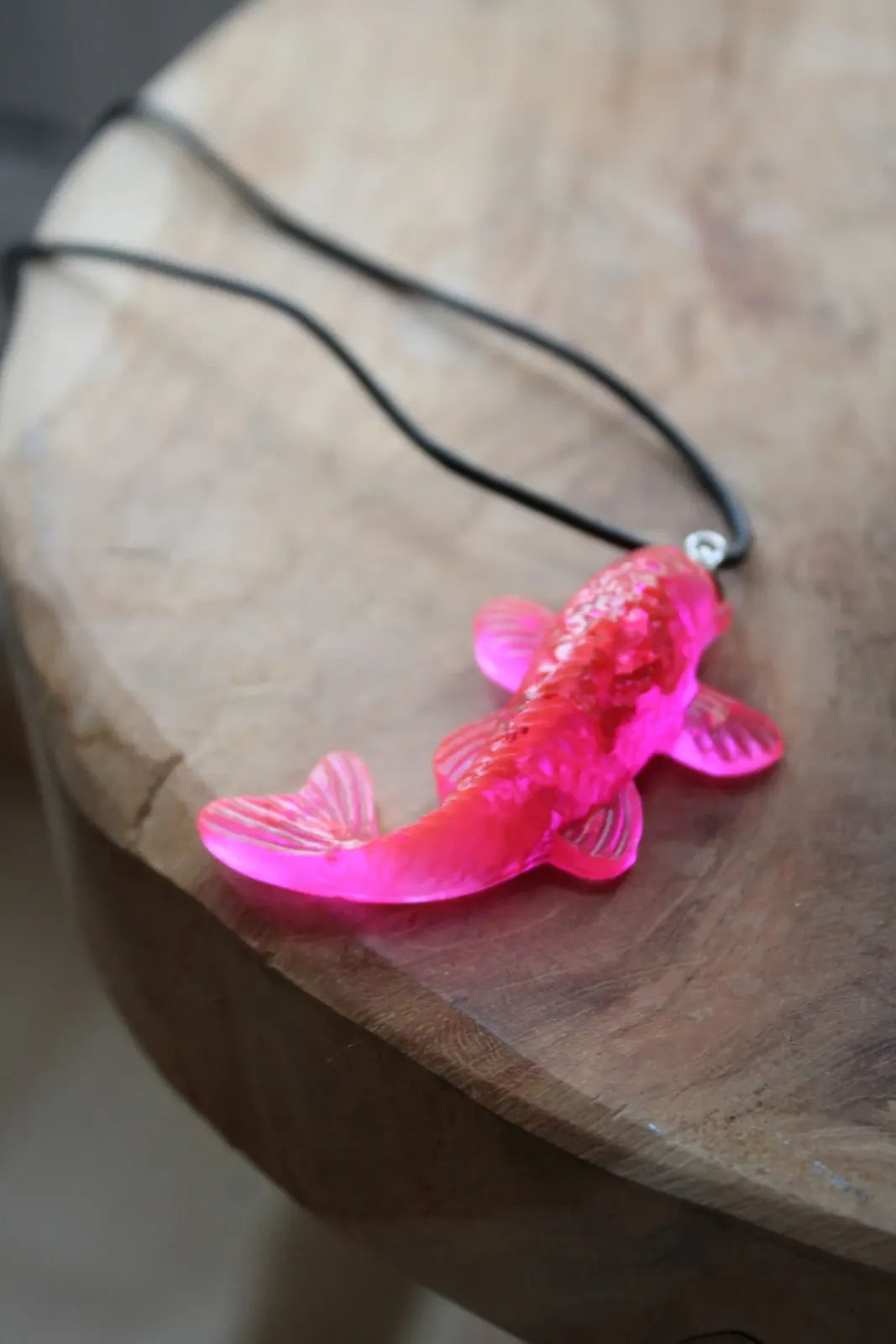 Figurine de poisson rose vif translucide avec un cordon noir, posée sur un socle en bois.