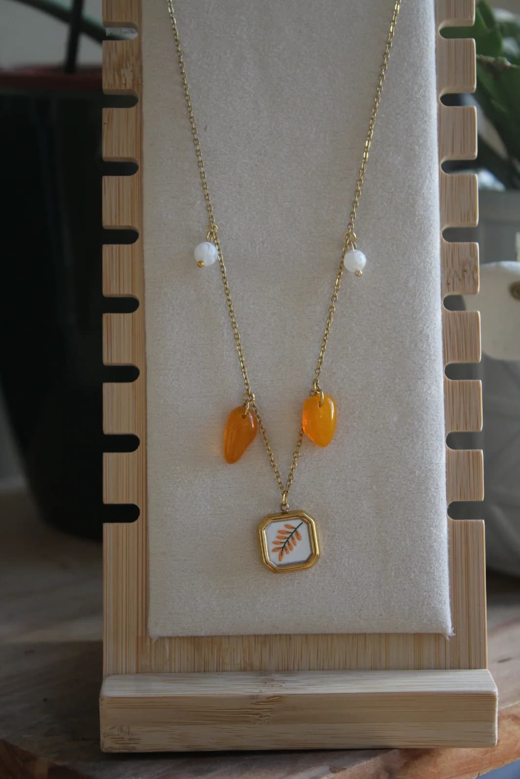 Collier doré à pendentif carré végétal et pampilles orange présenté sur un porte-bijou en bois