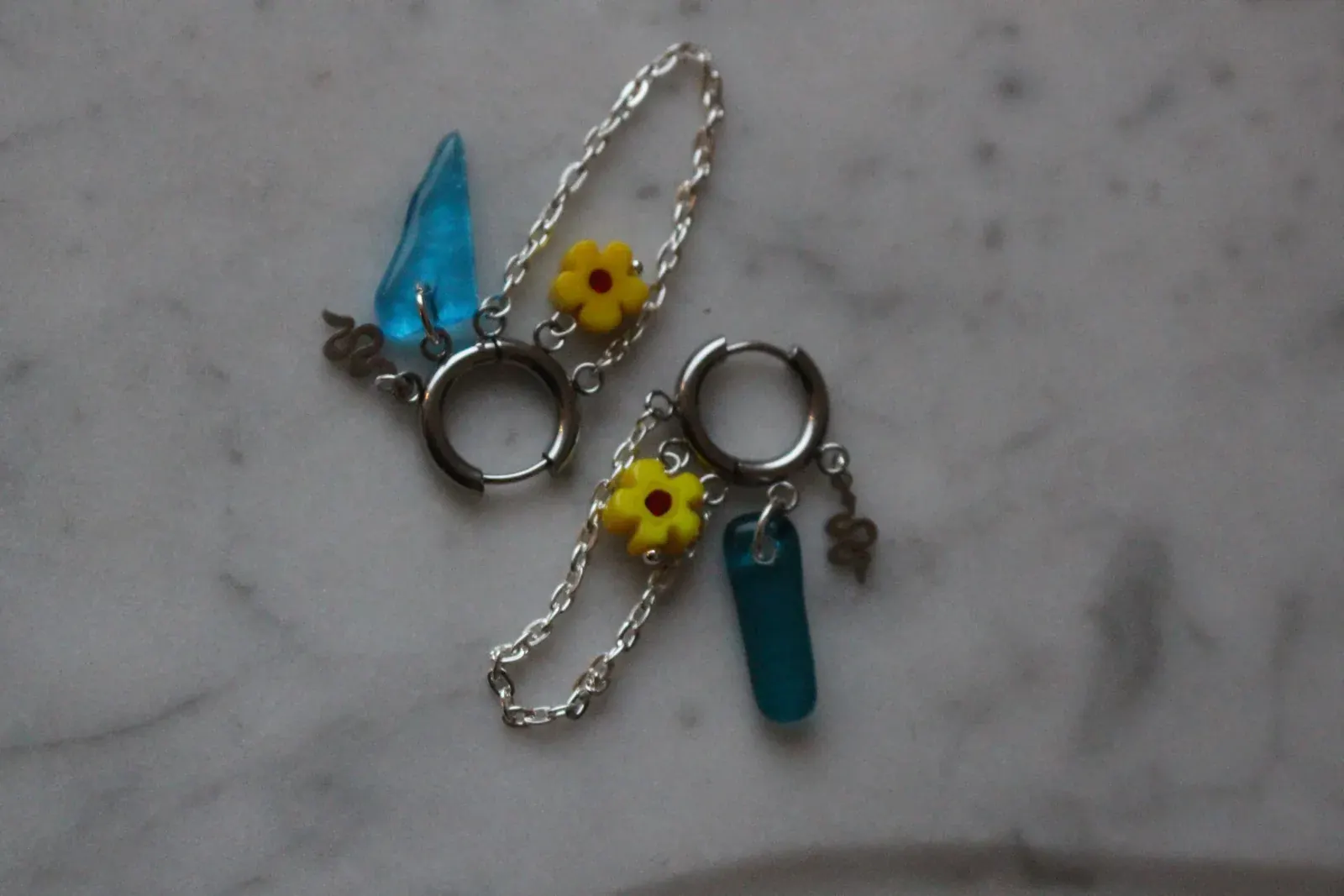 Créoles argentées avec pampilles en verre bleu lagon, fleurs jaunes et fines chaînettes sur fond marbré.