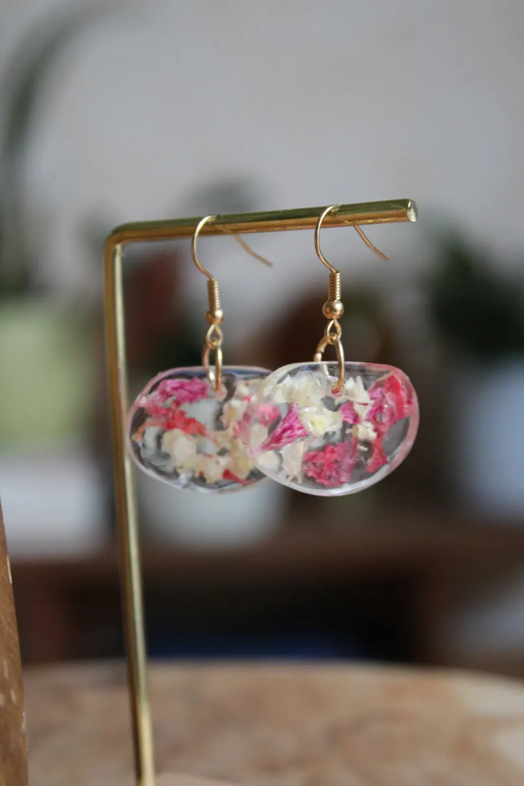 Boucles d'oreilles pendantes en résine transparente avec fleurs séchées roses et blanches sur un présentoir doré.