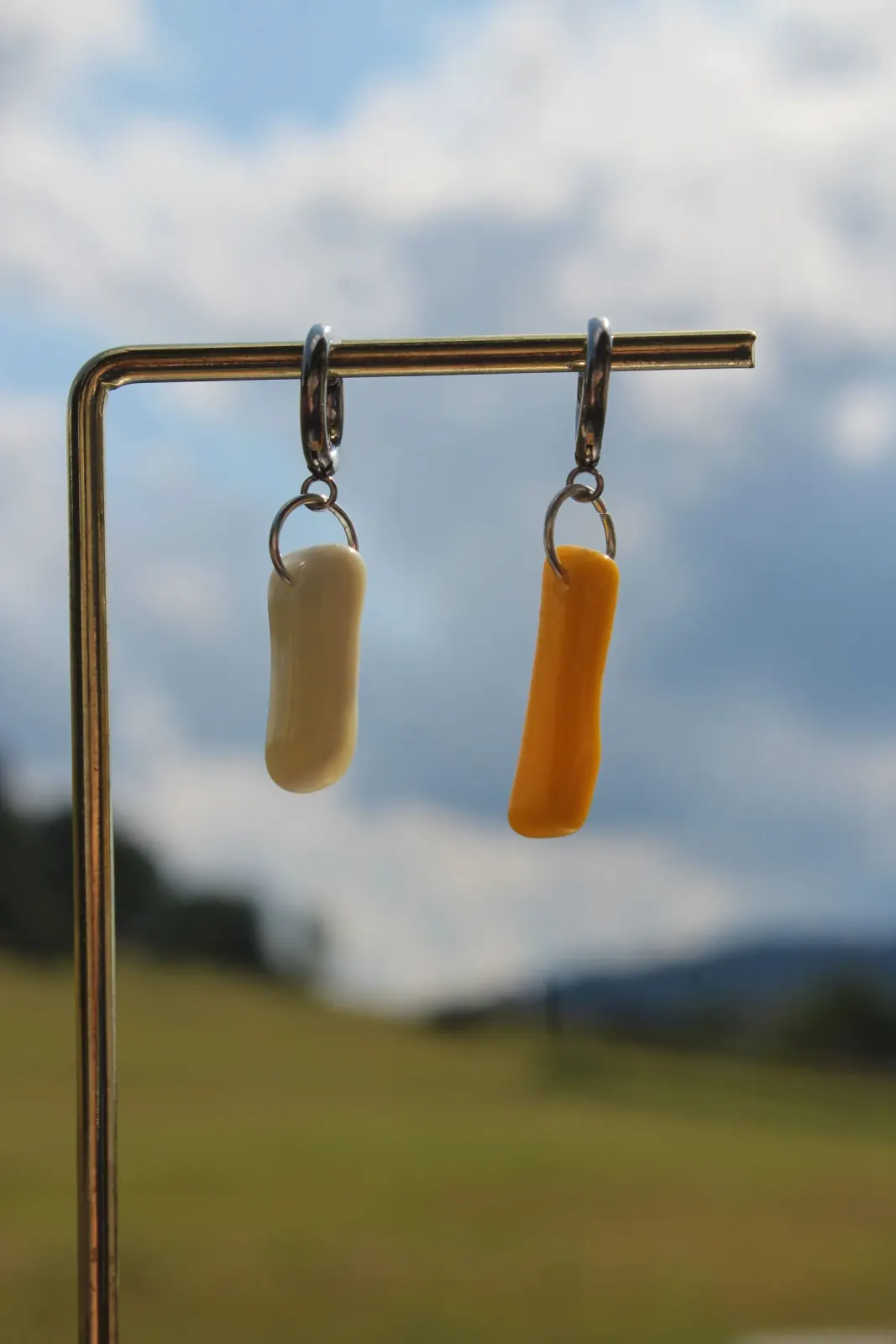 Boucles d'oreilles asymétriques en verre blanc et jaune suspendues sur un support, fond de ciel nuageux.