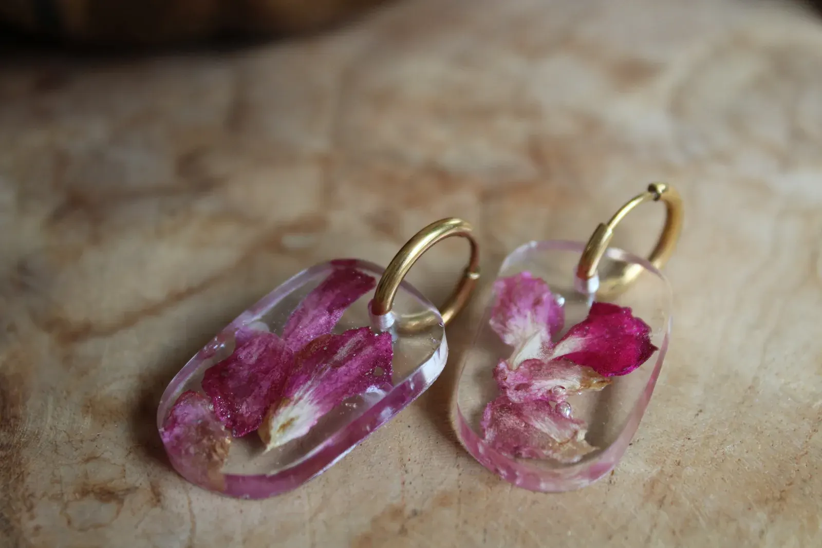 Boucles d'oreilles ovales transparentes avec pétales roses et anneaux dorés posées sur du bois.