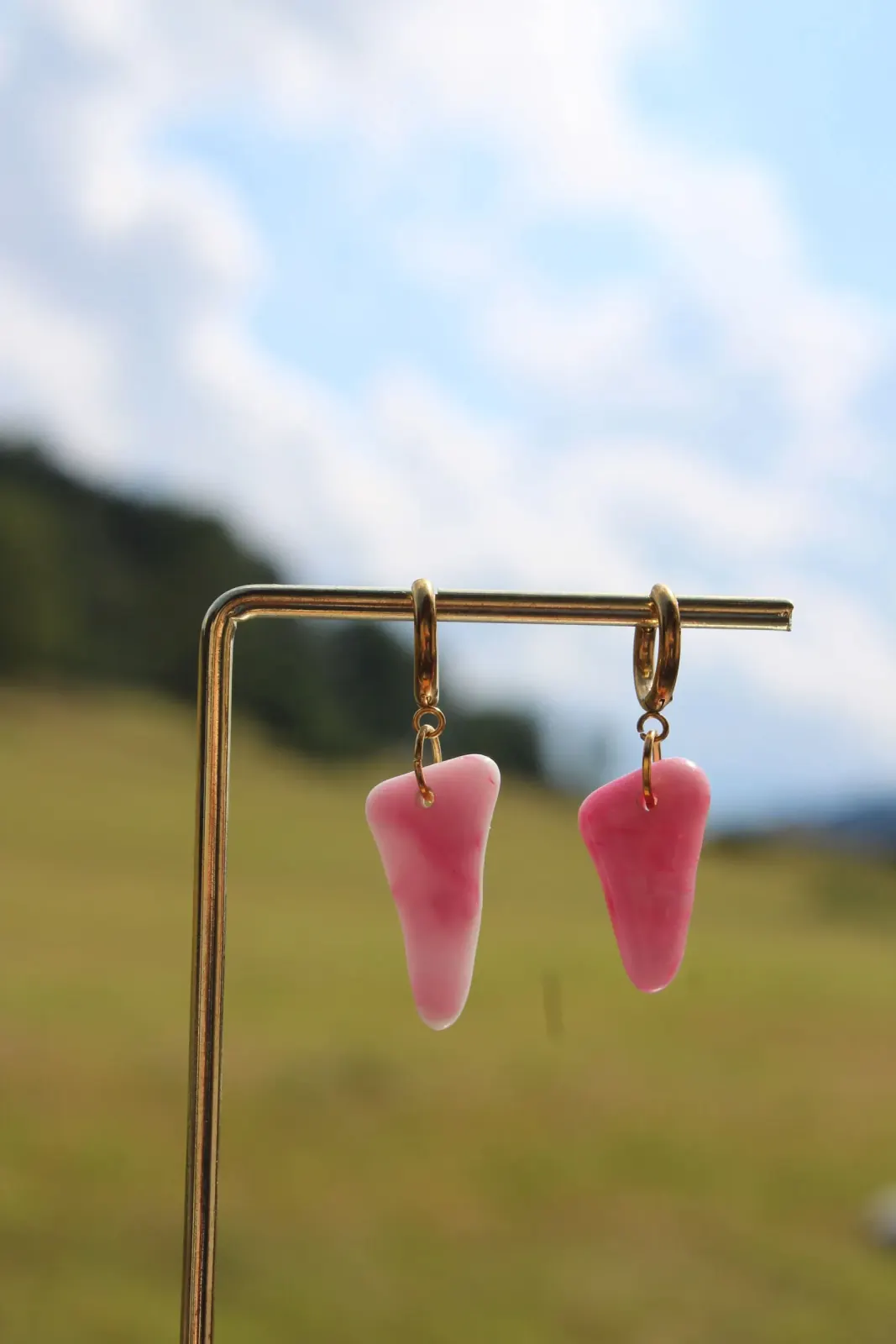 Boucles d'oreilles asymétriques en verre rose marbré suspendues sur un présentoir doré, fond de nature flou.