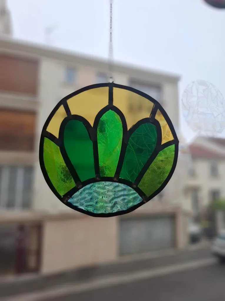 Vitrail rond suspendu au motif floral stylisé jaune, vert et bleu texturé devant une fenêtre.