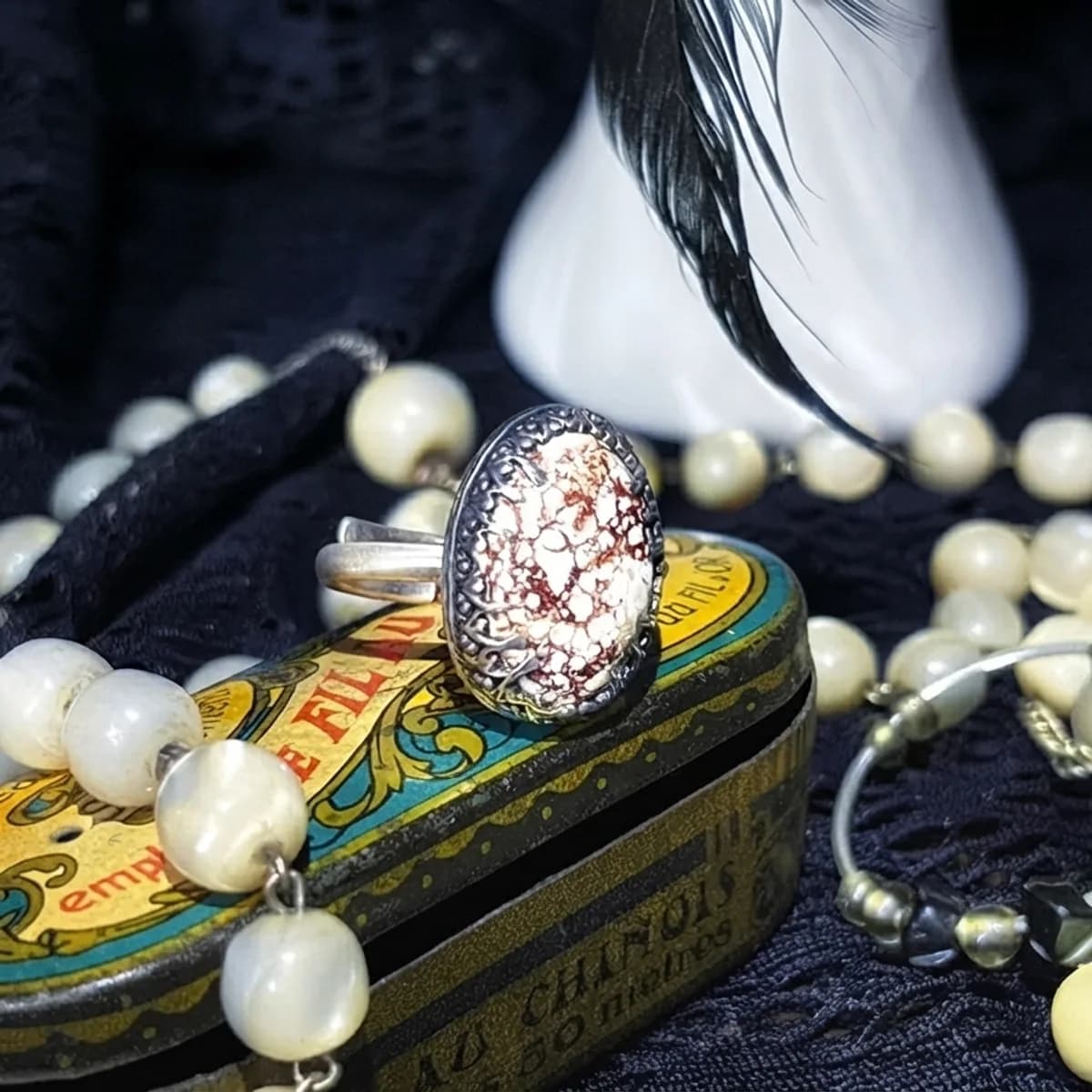 Bague en argent avec pierre ovale mouchetée posée sur une boîte vintage jaune et entourée de perles.