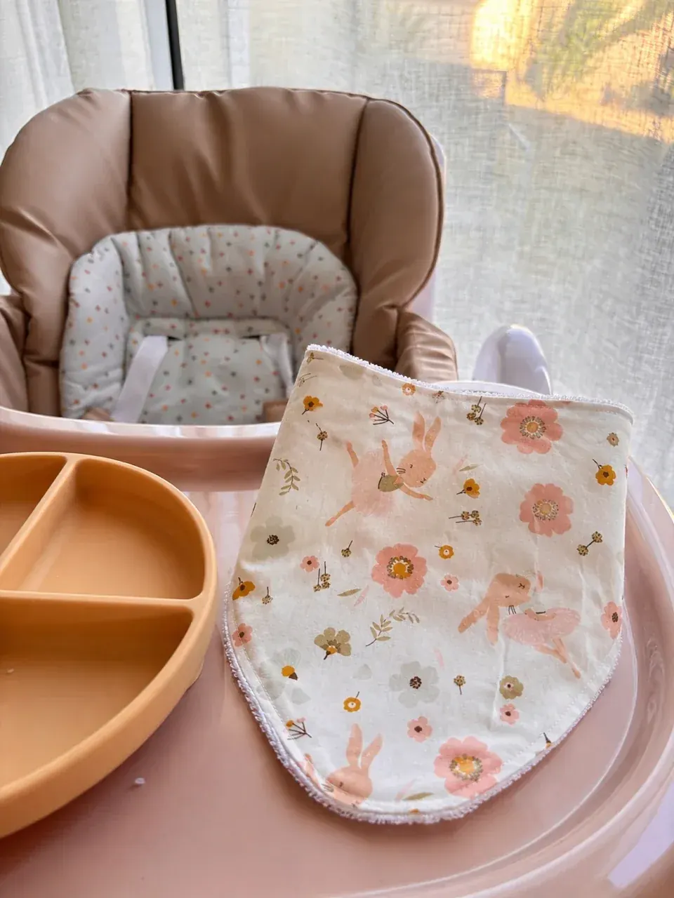Bavoir bandana écru aux motifs de fleurs roses et lapins, posé sur le plateau d'une chaise haute.