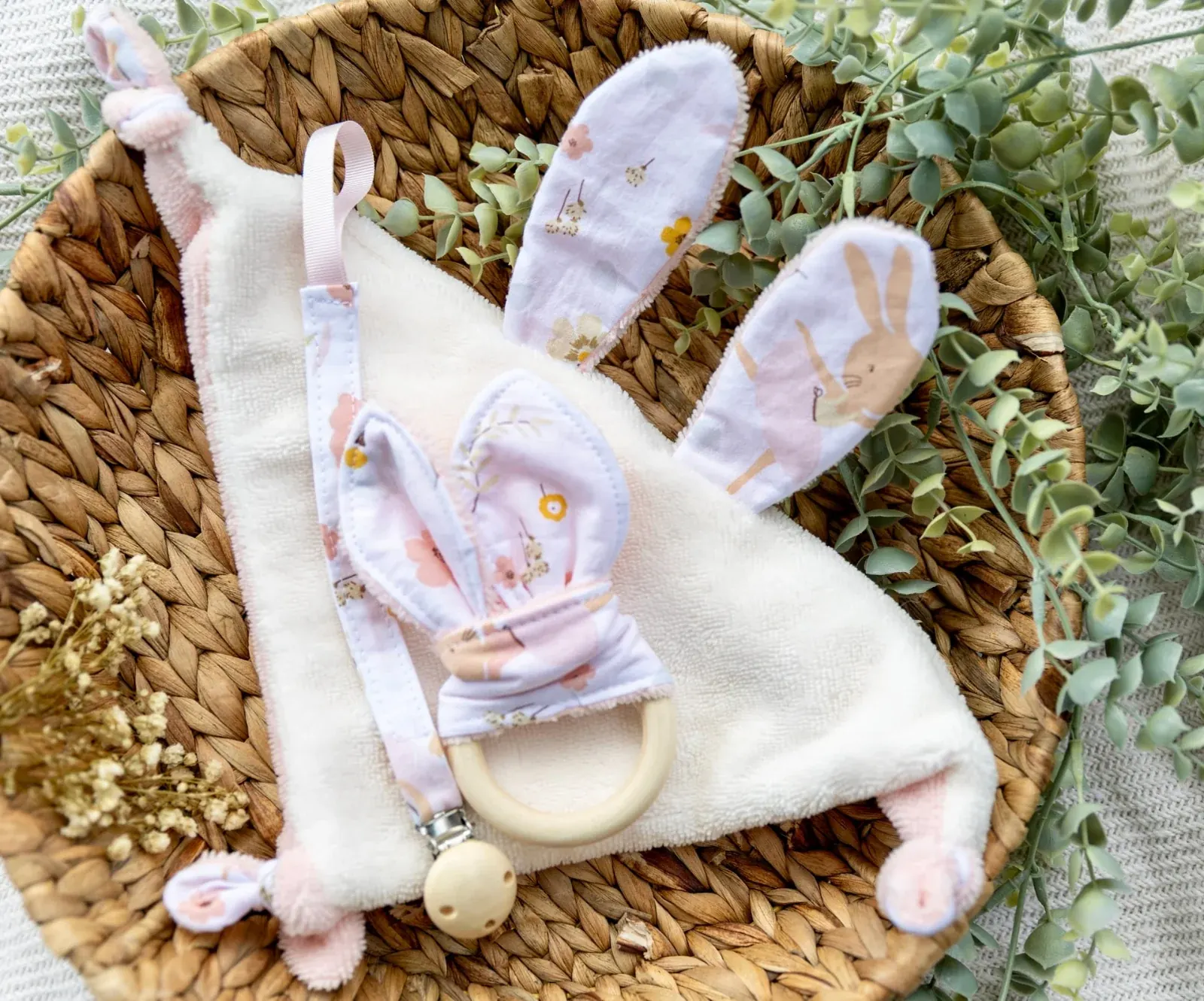 Doudou plat lapin, hochet en bois et attache-tétine fleuris présentés dans un panier en osier.