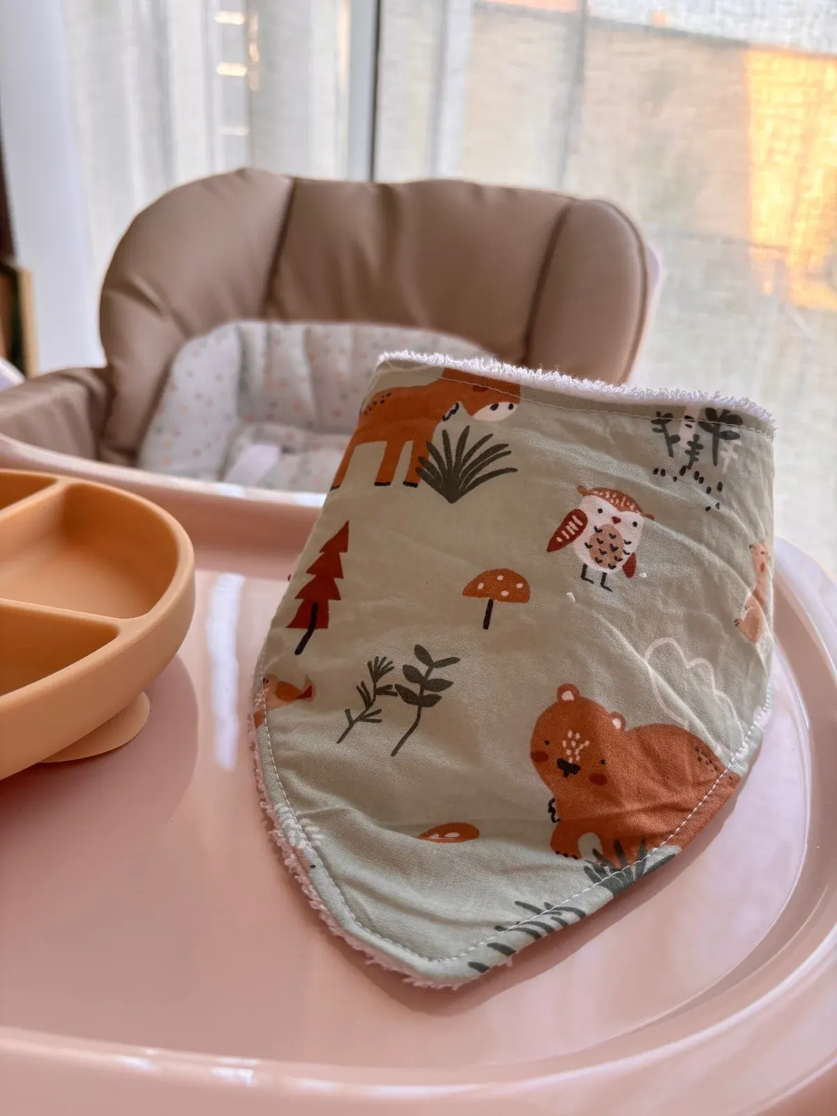 Bavoir bandana vert à motifs d'animaux de la forêt posé debout sur le plateau rose d'une chaise haute.