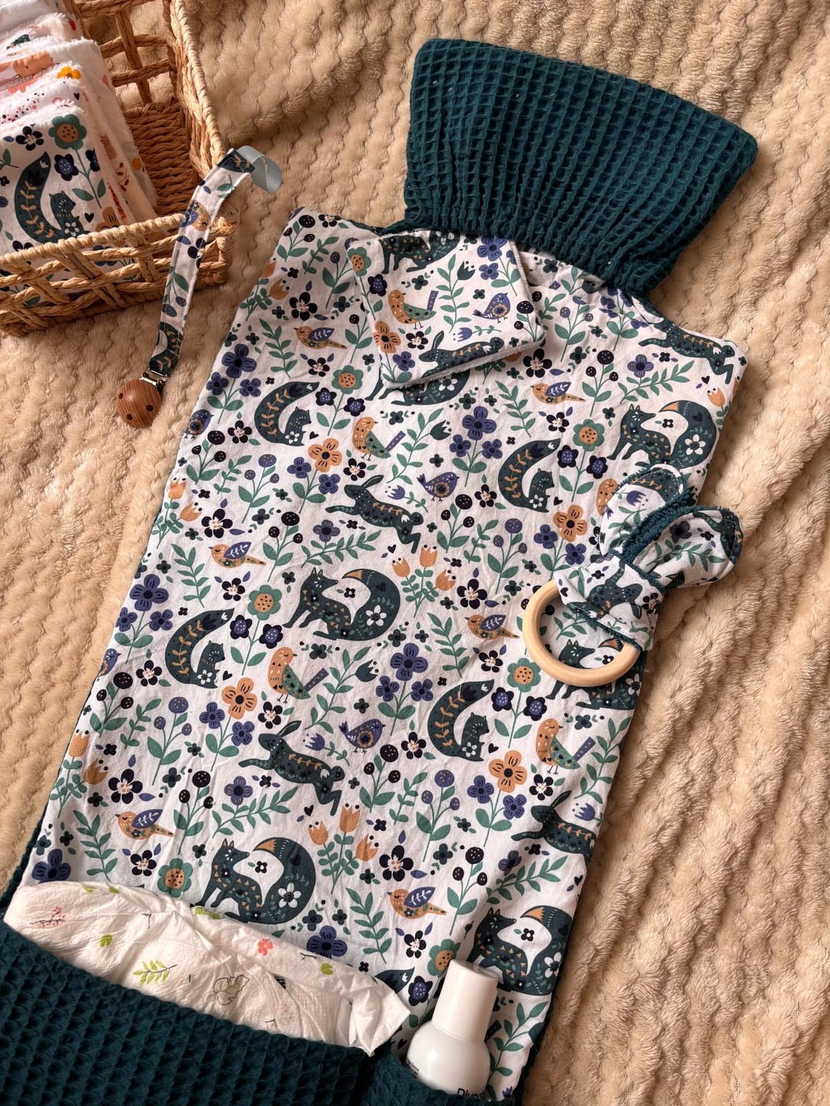 Tapis à langer déplié à motif forêt et tissu gaufré vert canard, posé sur une couverture beige.