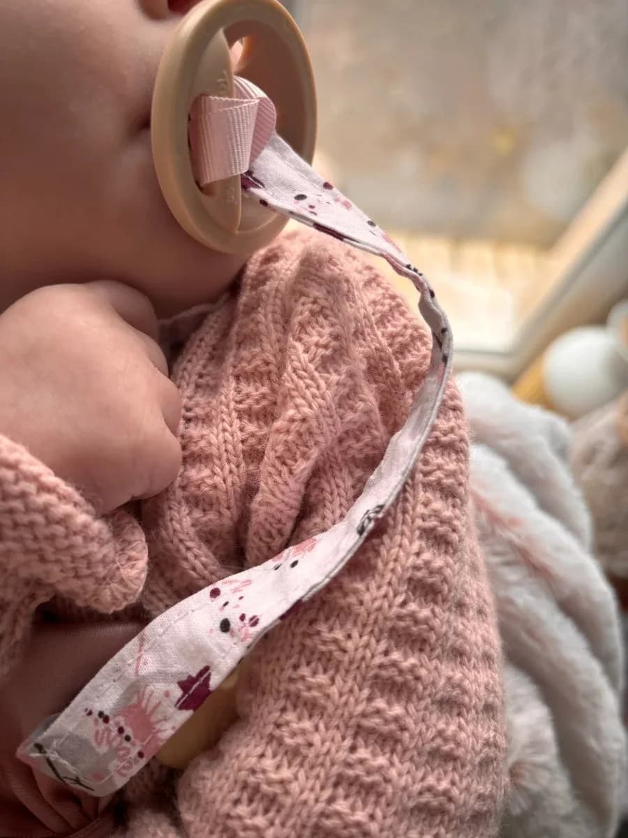 Bébé portant un pull en maille rose et une tétine reliée à une attache en tissu blanc à motifs.