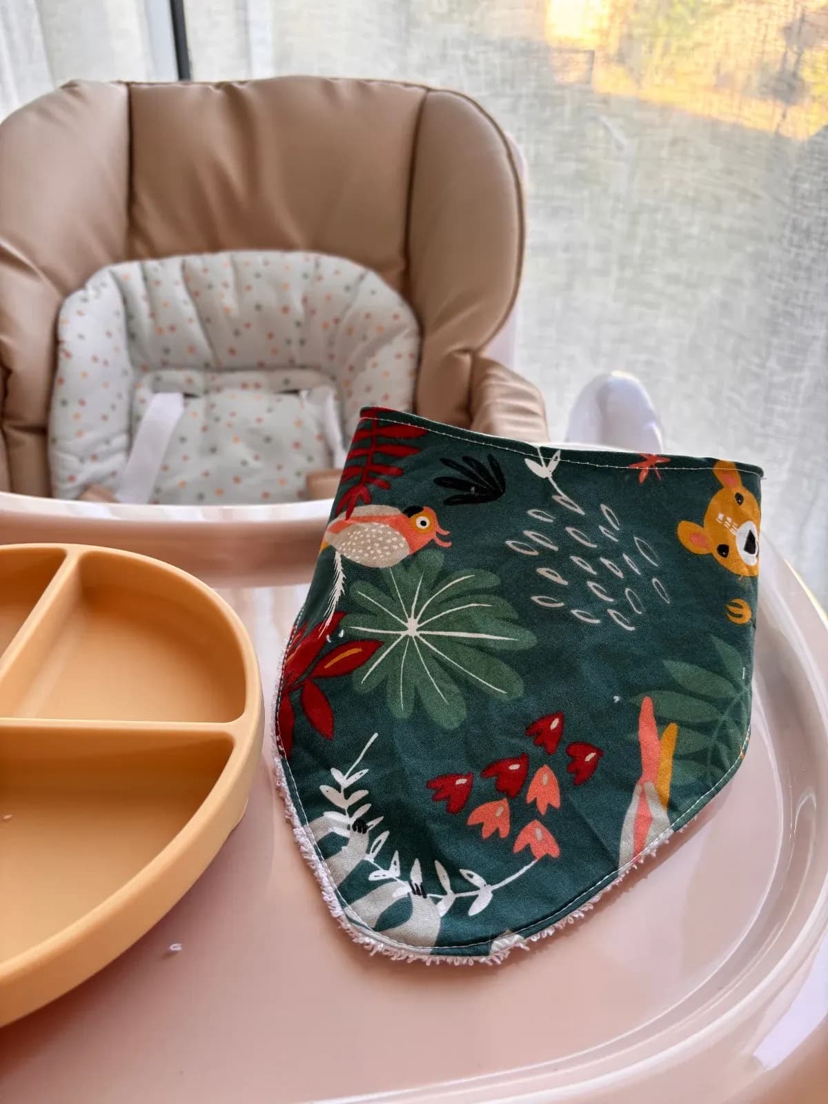 Bavoir bandana vert à motifs de feuilles et d'animaux posé sur une tablette de chaise haute rose.