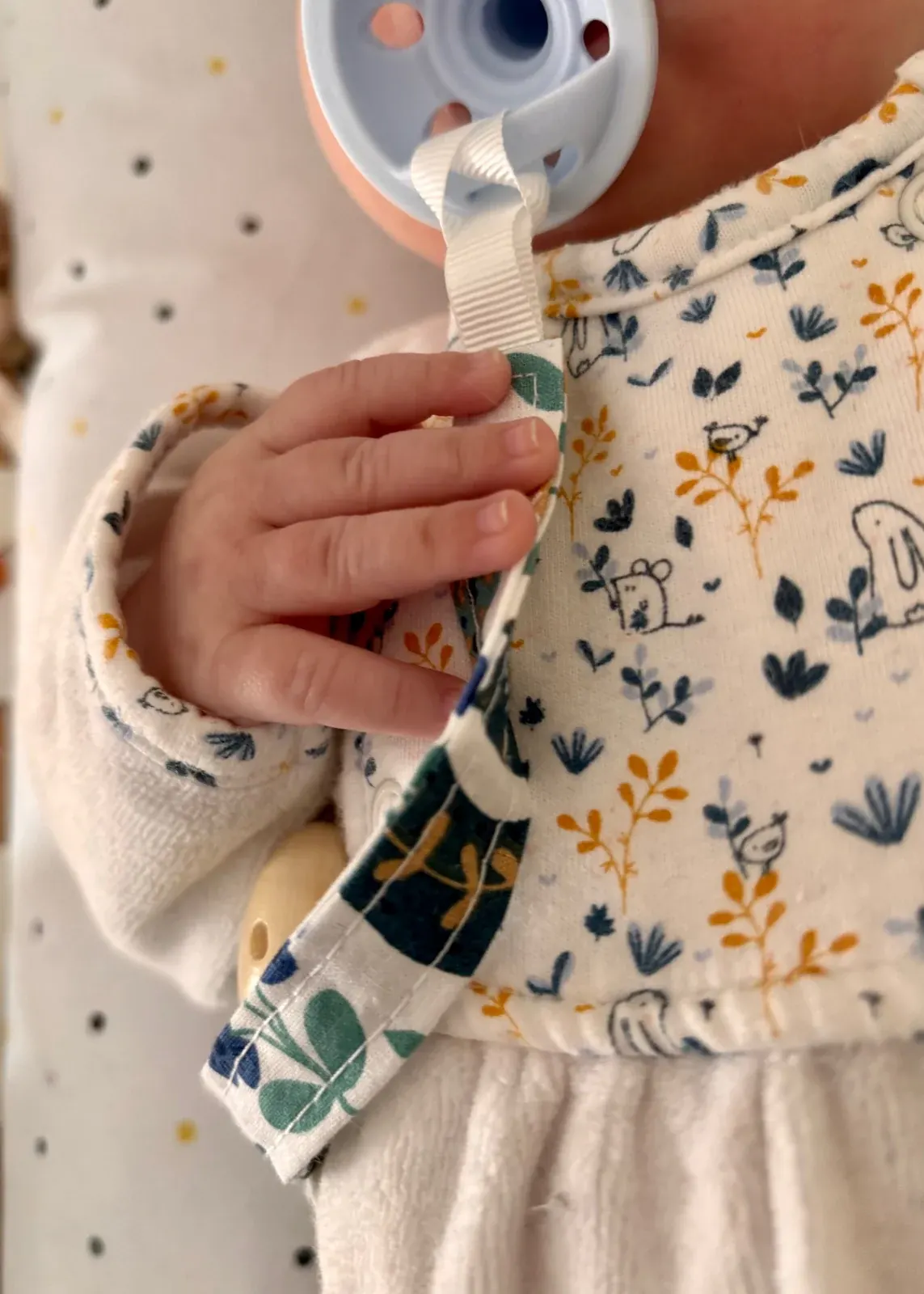 Main de bébé tenant une attache-tétine en tissu blanc à motifs de forêt et d'animaux.