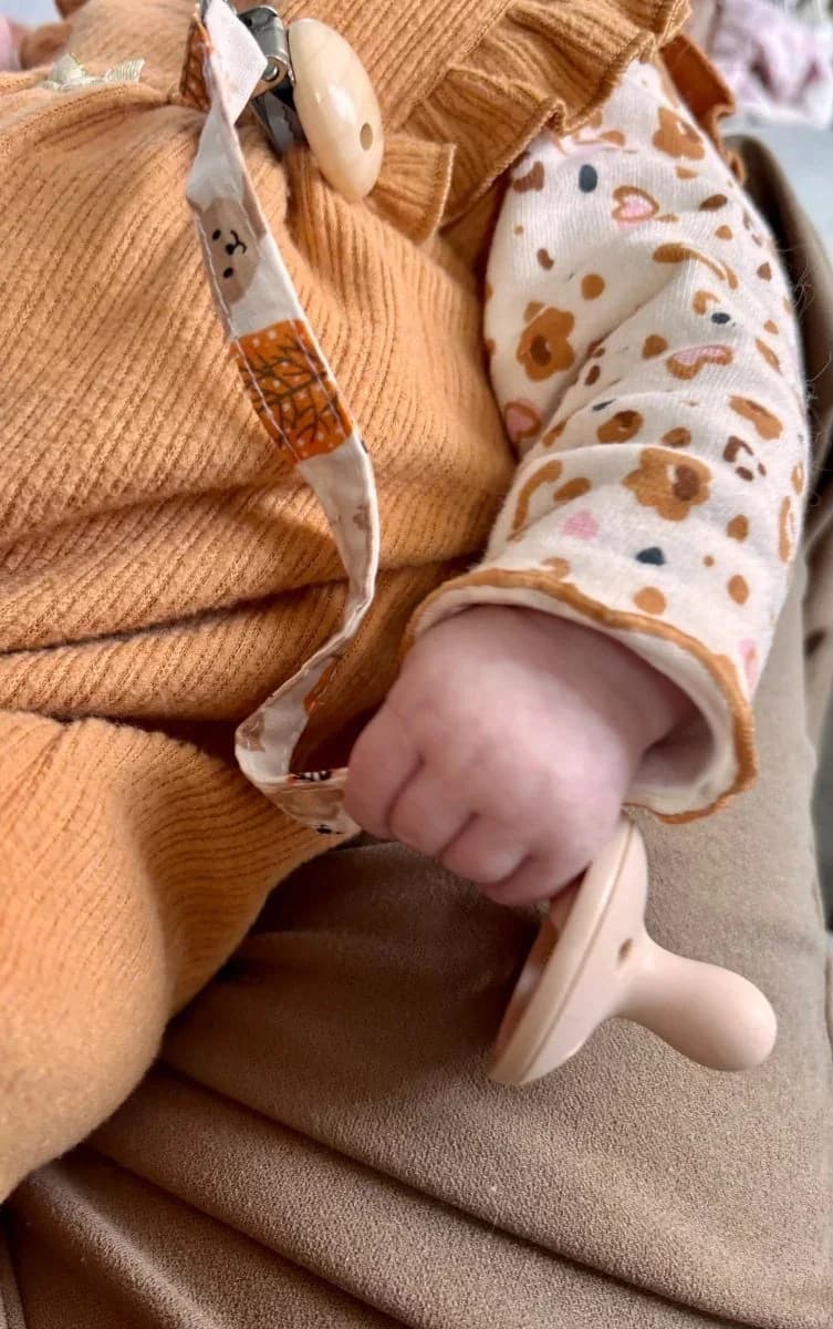 Attache-tétine en tissu blanc à motifs d'automne posée sur un plaid doux avec une tétine jaune.