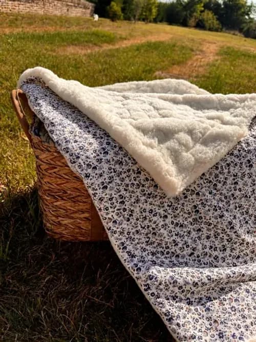 Couverture bébé à motifs floraux bleus et envers blanc polaire, posée sur un panier en osier dans l'herbe.