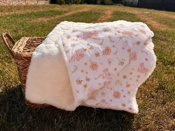 Couverture bébé blanche et fleurie posée sur un panier en osier dans l'herbe.