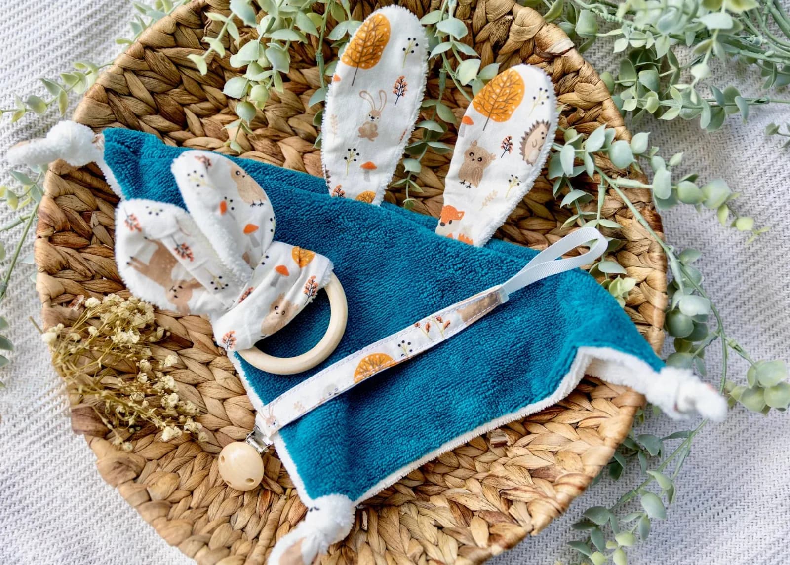 Doudou plat bleu canard, hochet oreilles de lapin et attache-tétine présentés dans un panier rond en osier.