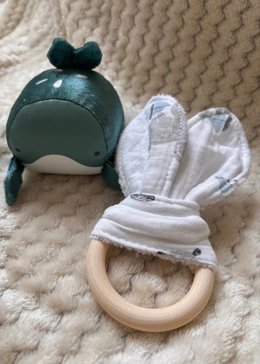 Anneau de dentition en bois et tissu blanc à motifs de bateaux, à côté d'une petite peluche baleine verte.