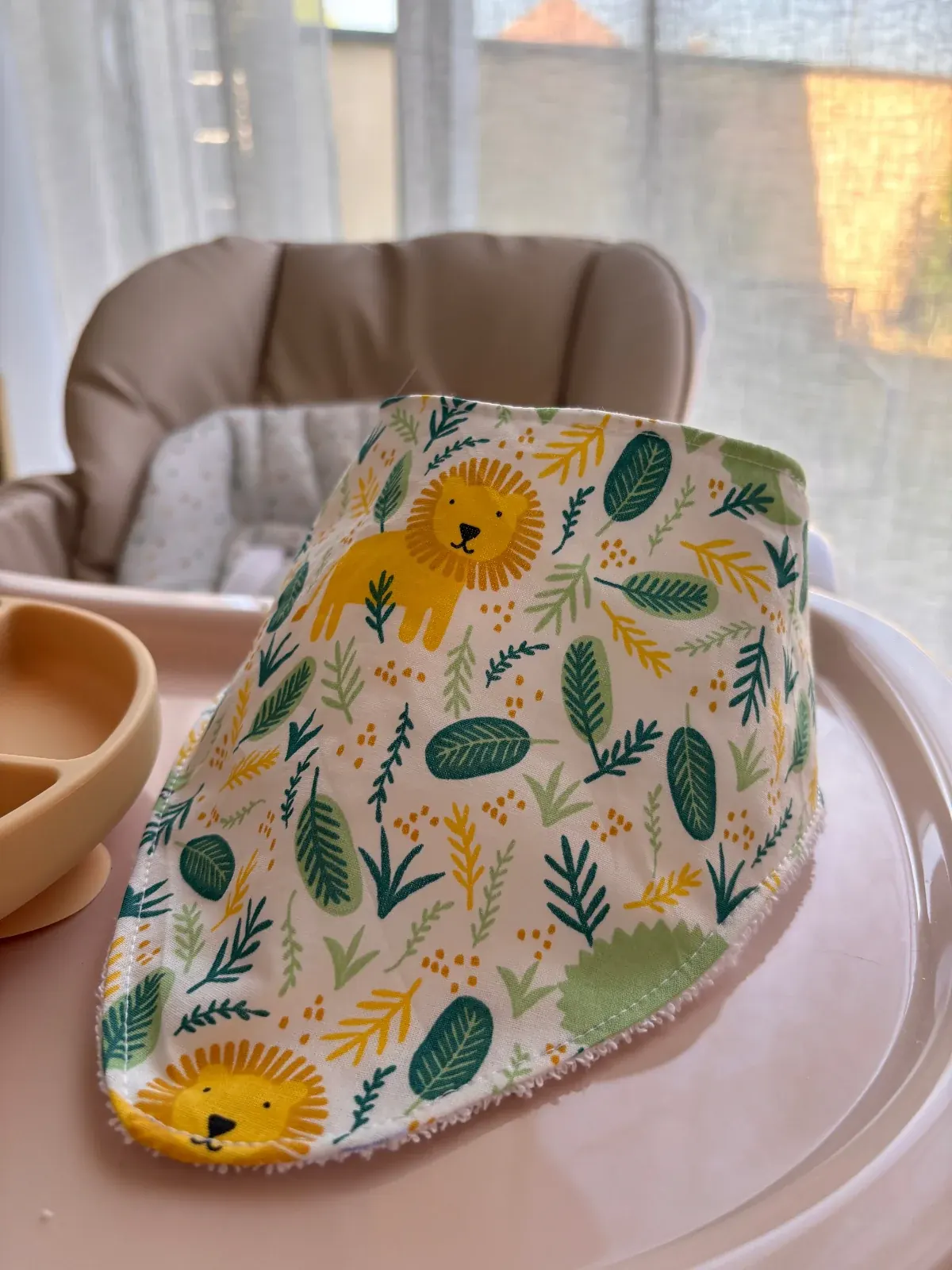 Bavoir bandana blanc à motifs de lions jaunes et feuilles vertes, posé sur une tablette de chaise haute.