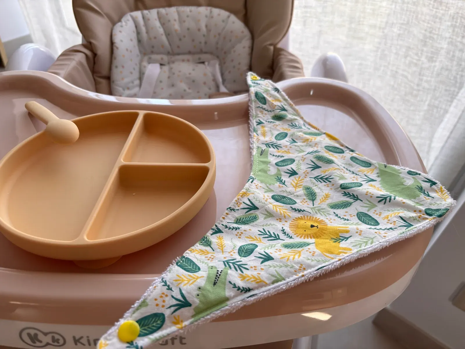Bavoir bandana déplié avec motifs jungle et bouton pression jaune, à côté d'une assiette beige.
