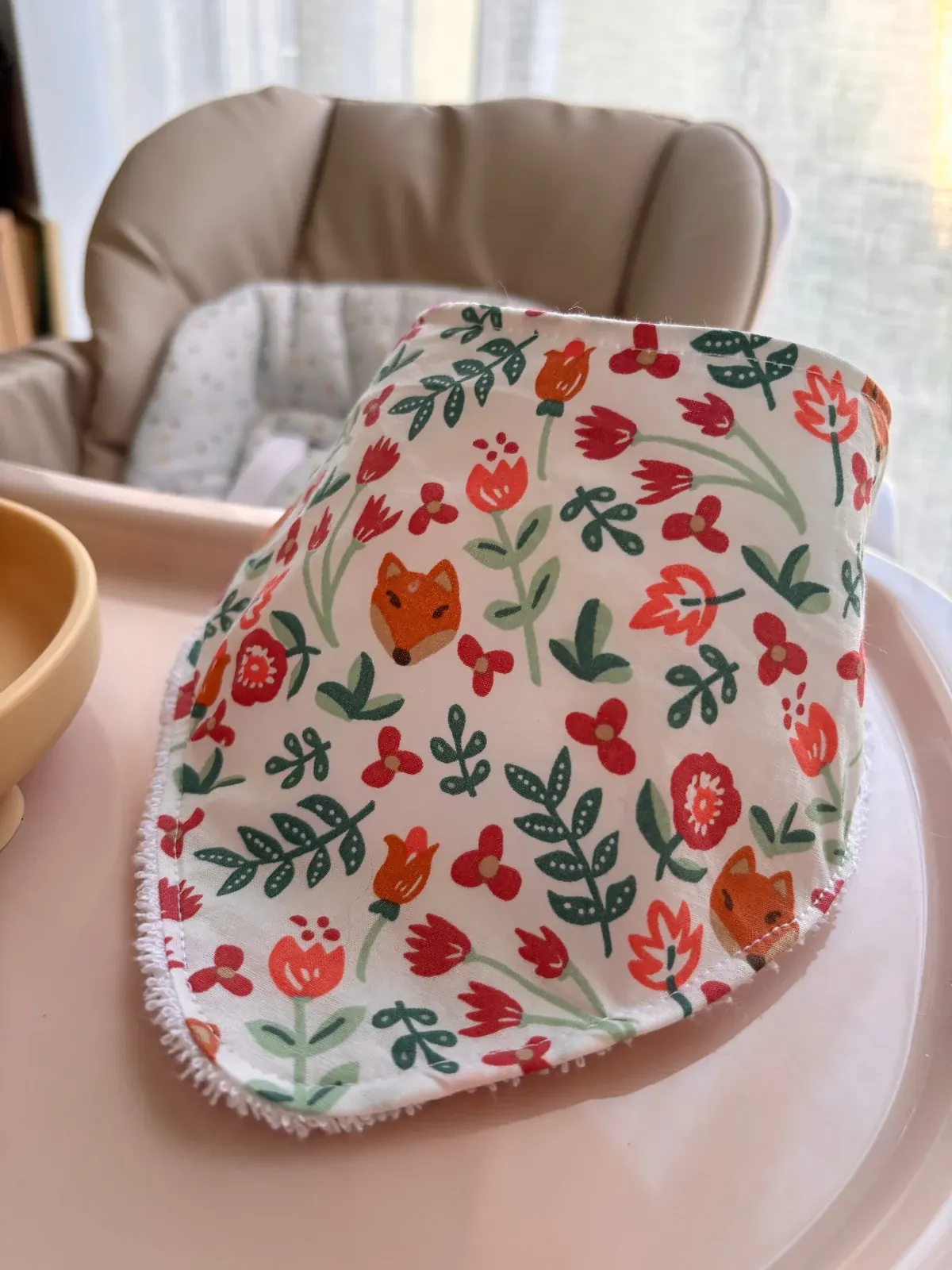 Bavoir bandana blanc à motifs de renards et fleurs orange et rouge, posé sur une tablette de chaise haute.