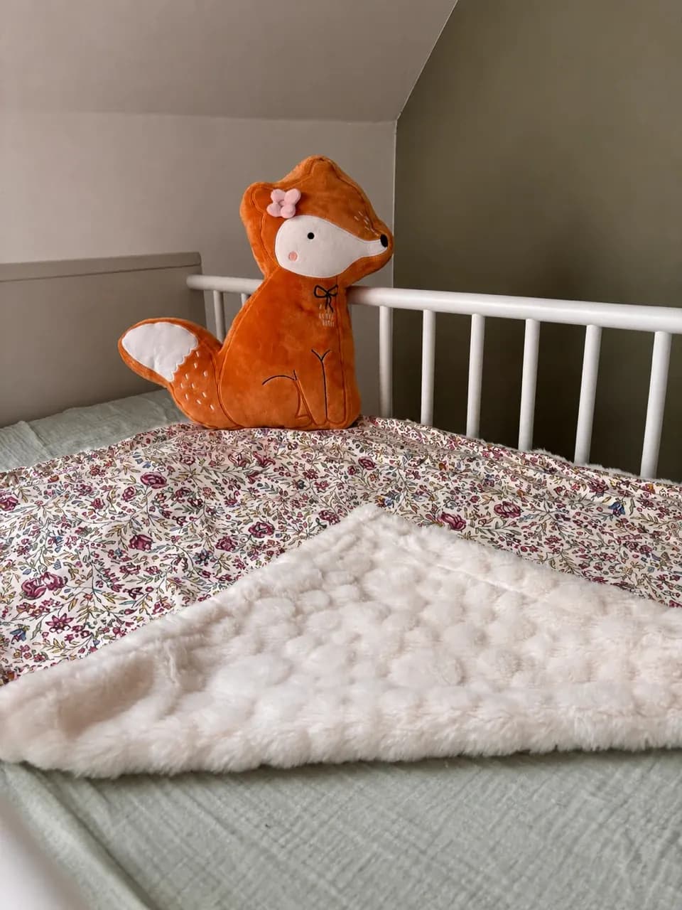 Couverture bébé à motifs floraux et envers polaire blanc posée dans un lit avec une peluche renard.
