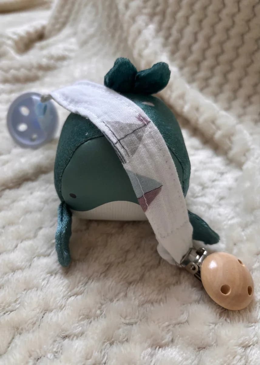 Attache-tétine en tissu blanc à motifs géométriques avec clip en bois, posée sur une peluche baleine verte.