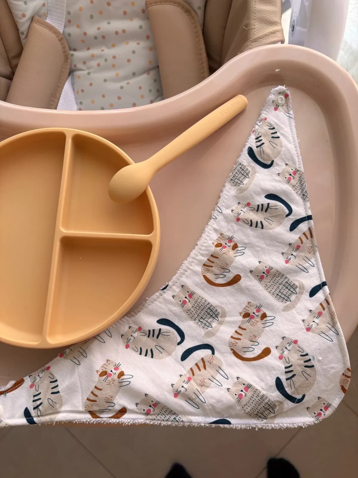 Bavoir triangulaire déplié avec motifs de chats, assiette compartimentée et cuillère beige.