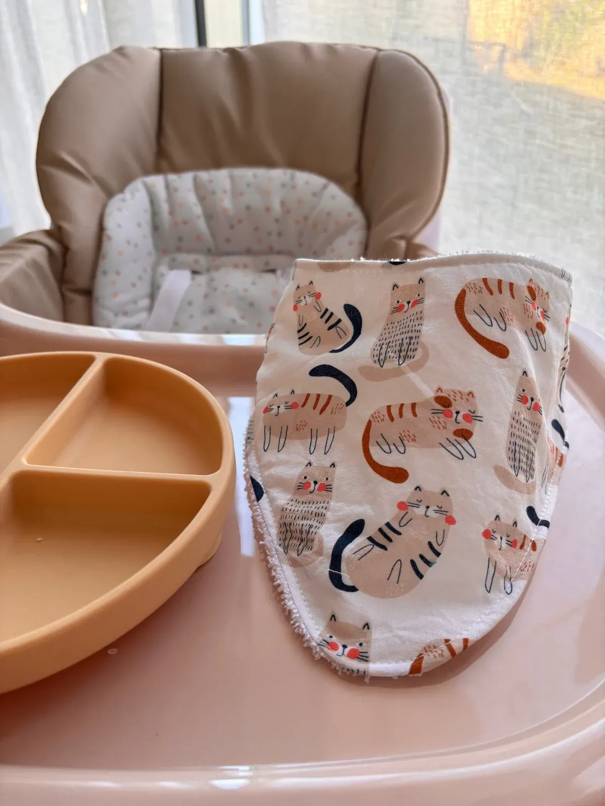 Bavoir bandana blanc à motifs de petits chats posé sur une tablette de chaise haute beige.