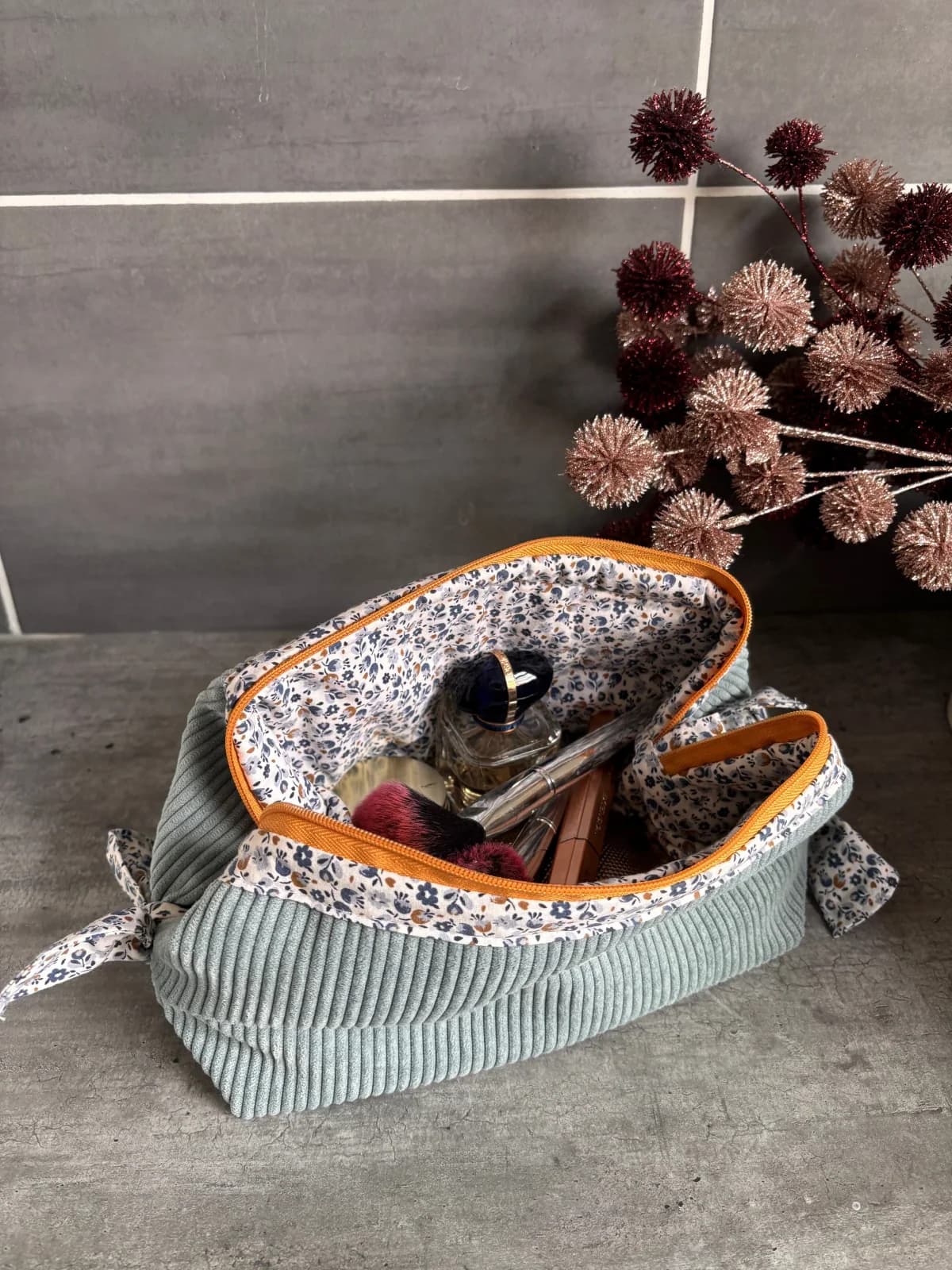 Trousse en velours gris-bleu ouverte révélant une doublure fleurie et des produits de maquillage.