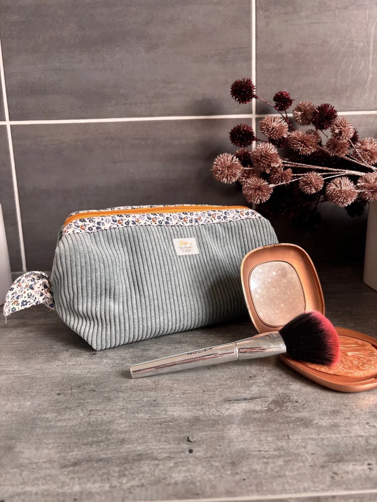 Trousse en velours côtelé gris-bleu fermée, posée à côté d'un pinceau et d'une poudre de maquillage.