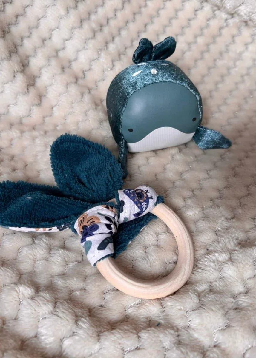 Gros plan sur l'anneau en bois et le tissu vert texturé, posés sur une couverture beige avec une petite baleine.
