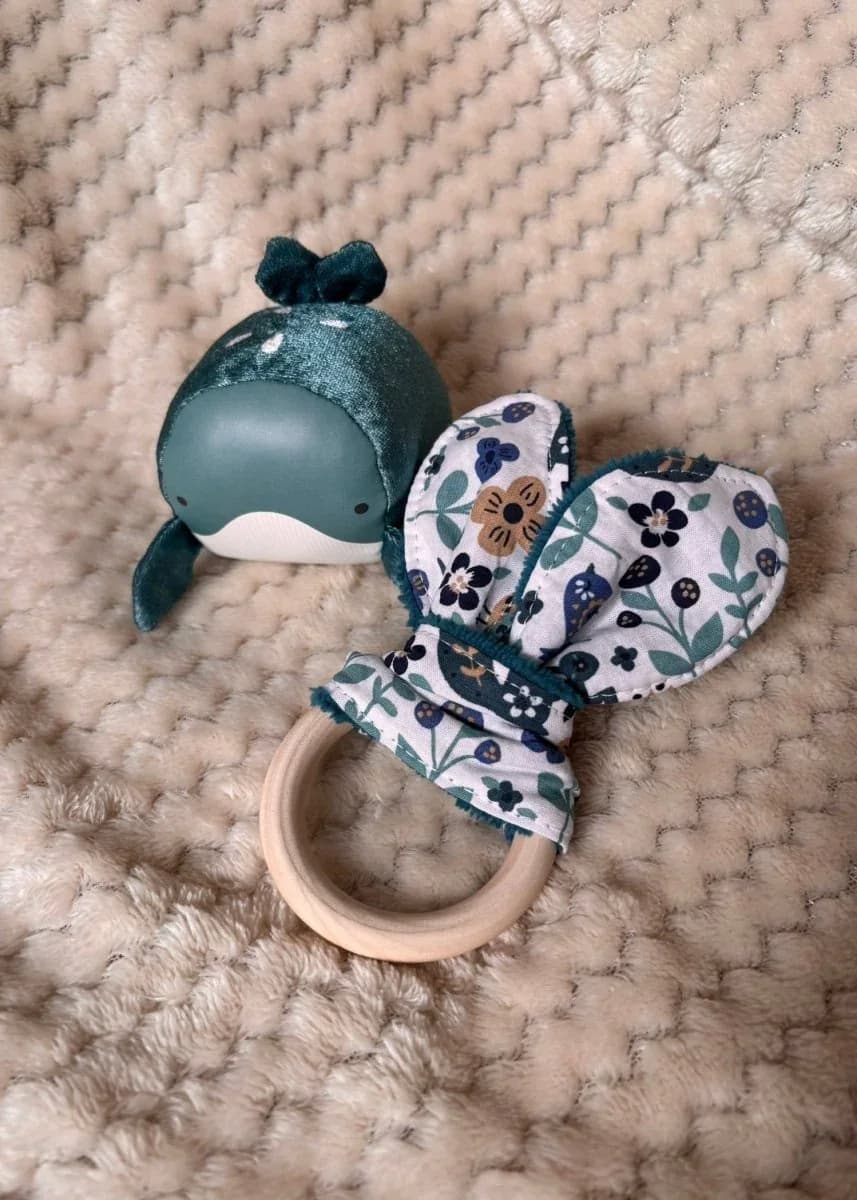 Anneau de dentition en bois avec tissu floral à oreilles de lapin à côté d'une petite baleine verte.