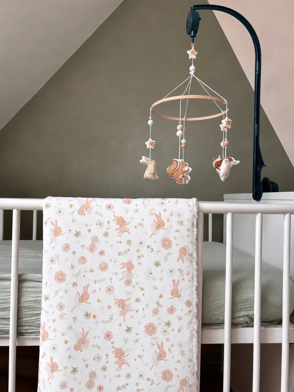 Couverture blanche à motifs de lapins roses suspendue sur un lit de bébé blanc avec un mobile en bois.