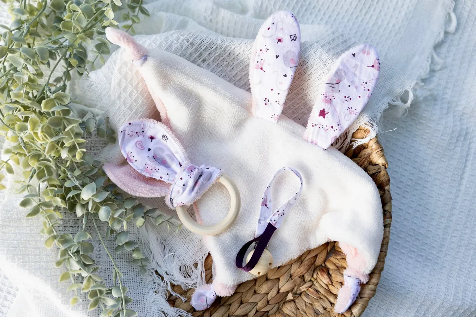 Doudou plat blanc, hochet en bois et attache-tétine assortis dans un panier en osier avec feuillage.