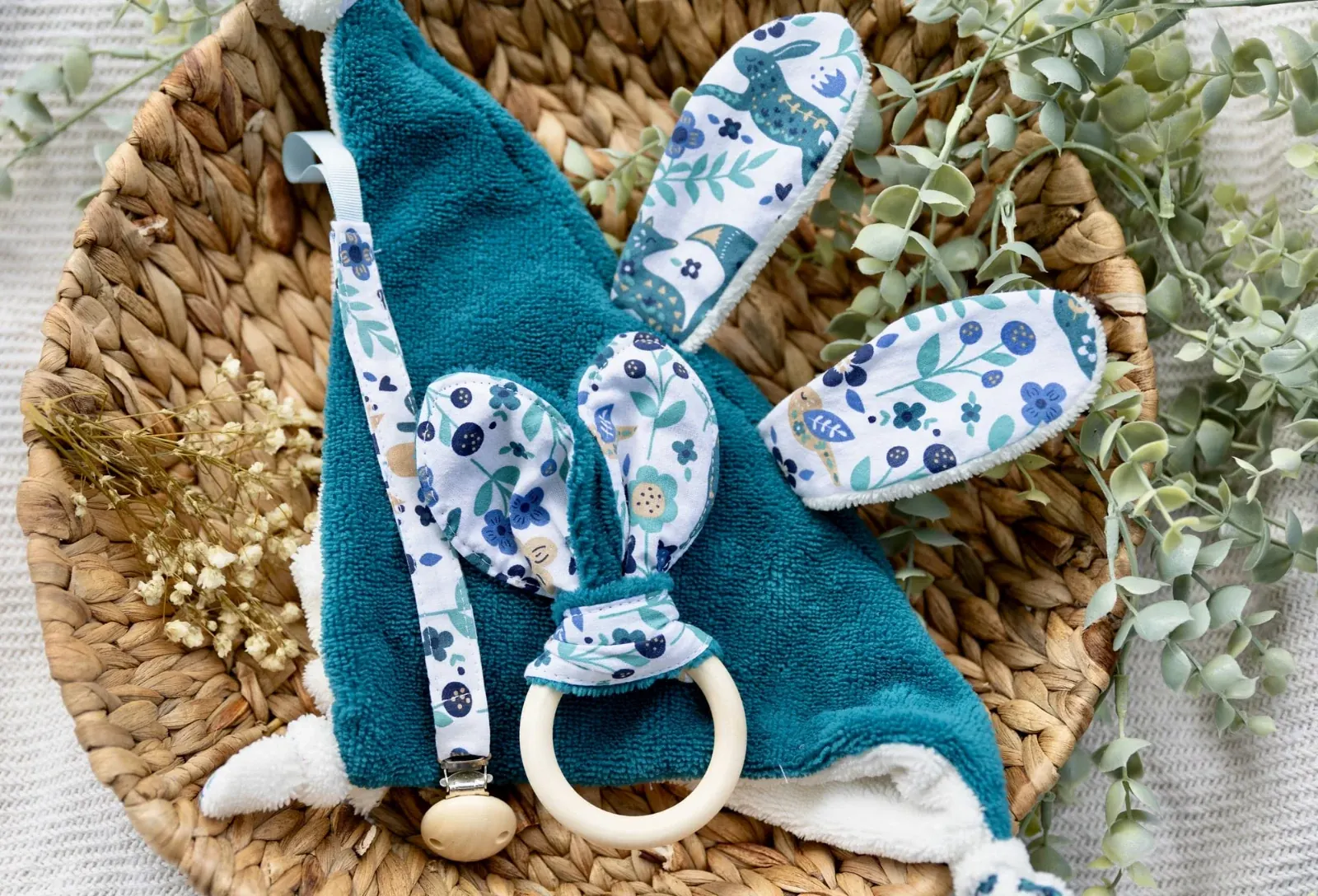 Doudou plat bleu canard, attache-tétine et anneau de dentition dans un panier en osier.