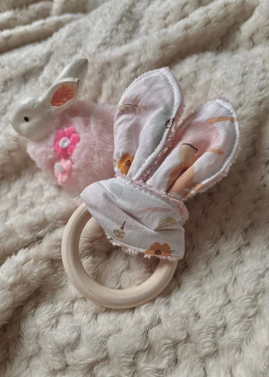 Anneau de dentition en bois avec oreilles en tissu fleuri à côté d'un petit lapin rose en peluche.