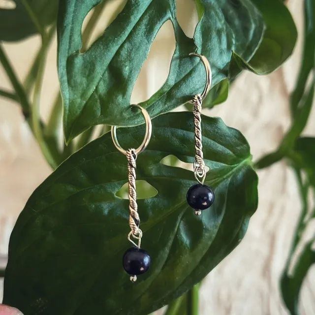 Boucles d'oreilles créoles argentées avec tiges torsadées et perles sombres, suspendues à une feuille verte.