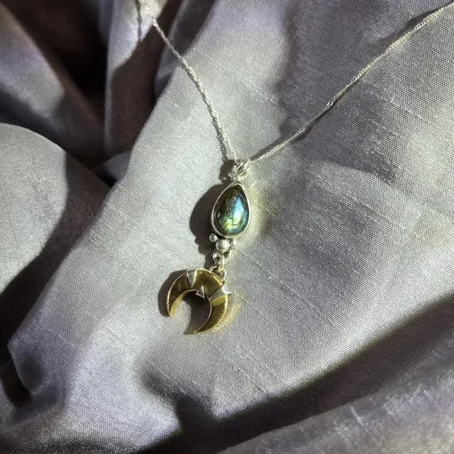Pendentif lune en laiton et labradorite sur sa chaîne en argent, posé sur un tissu satiné gris.