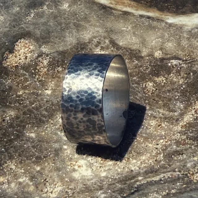 Large anneau en argent martelé et patiné posé à la verticale sur une roche texturée.