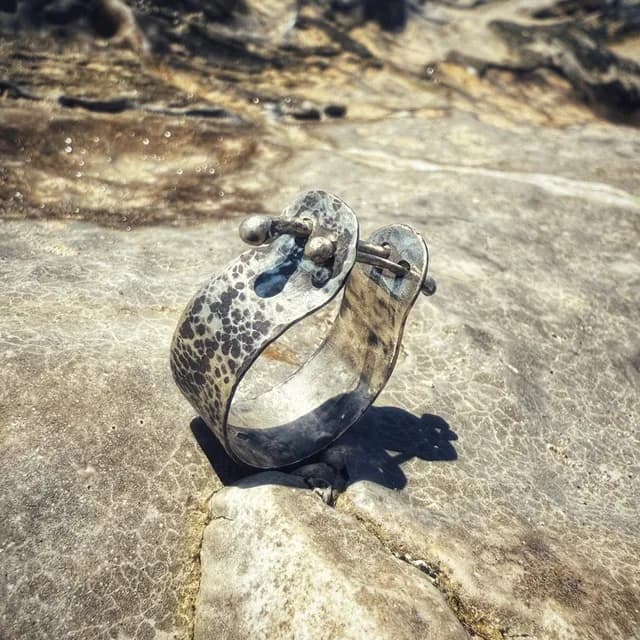 Bague en métal argenté texturé avec tiges mobiles, posée sur une roche grise ensoleillée.