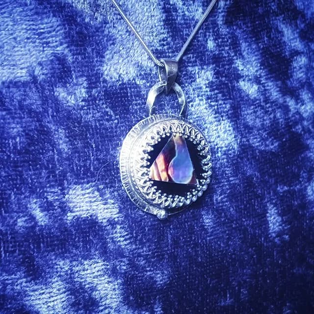 Pendentif rond en argent avec triangle en abalone irisé, présenté sur un tissu bleu texturé.