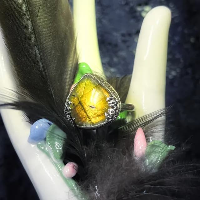 Bague en argent avec pierre triangulaire aux reflets jaunes et dorés, posée sur un support blanc avec plumes.