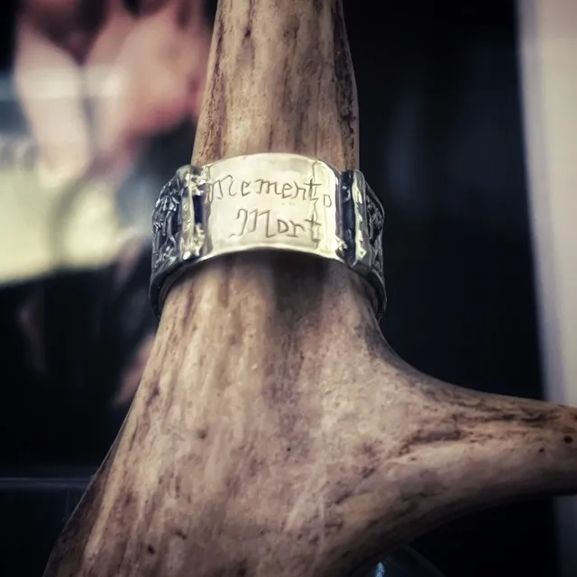 Bague en argent gravée Memento Mori présentée sur un support en bois ramifié.