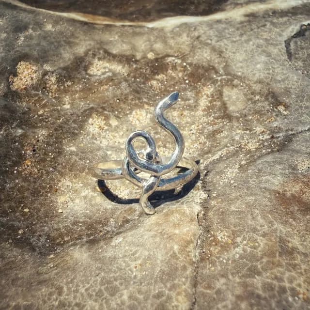 Bague en argent aux formes ondulées et entrelacées, posée sur une pierre beige texturée.