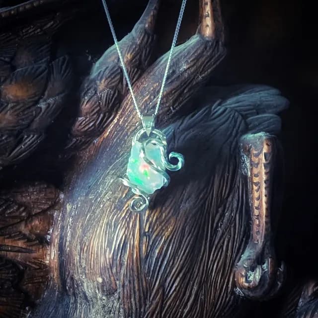 Pendentif en opale aux reflets verts et bleus, monté sur fil d'argent, devant une sculpture en bois.