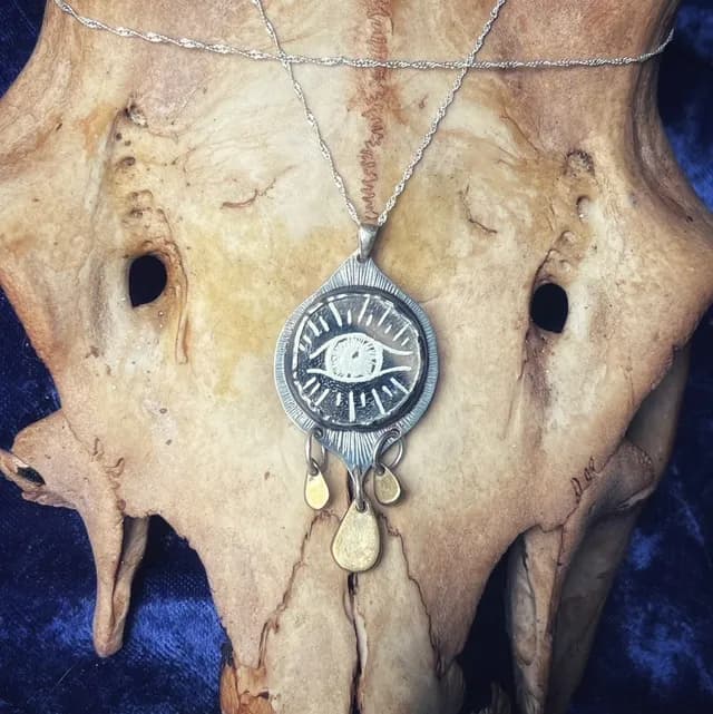 Pendentif œil gravé en argent et laiton posé sur un crâne d'animal, fond en velours bleu.