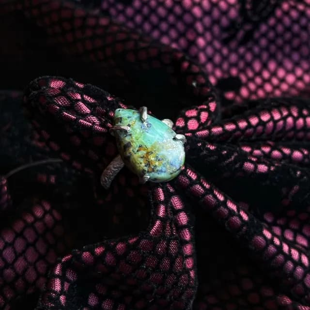 Bague en argent avec une pierre en forme de goutte verte et bleue, posée sur un tissu noir et rose.
