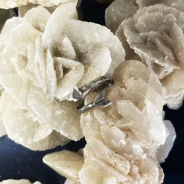 Bague ajustable en argent en forme de flèche, posée sur une rose des sables beige.