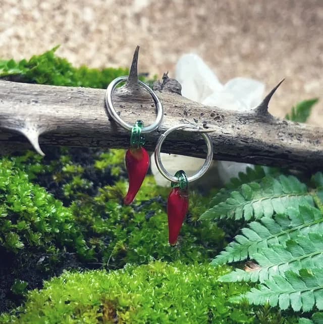 Créoles argentées avec pendentifs piments rouges en verre, suspendues à une branche épineuse sur de la mousse.