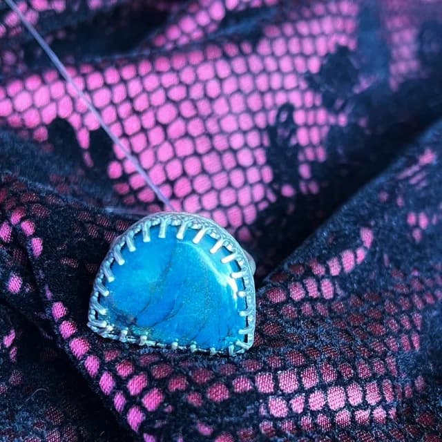 Bague argentée avec une pierre bleue en demi-lune, posée sur un tissu en dentelle noire et rose.